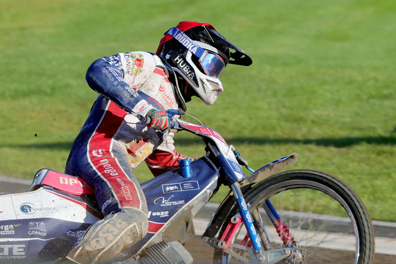 Zawodnik żużlowy ubrany w kombinezon oraz kask jedzie motocyklem po torze żużlowym, skupiony na wyścigu, dynamiczna pozycja podkreśla szybkość i intensywność rywalizacji sportowej.