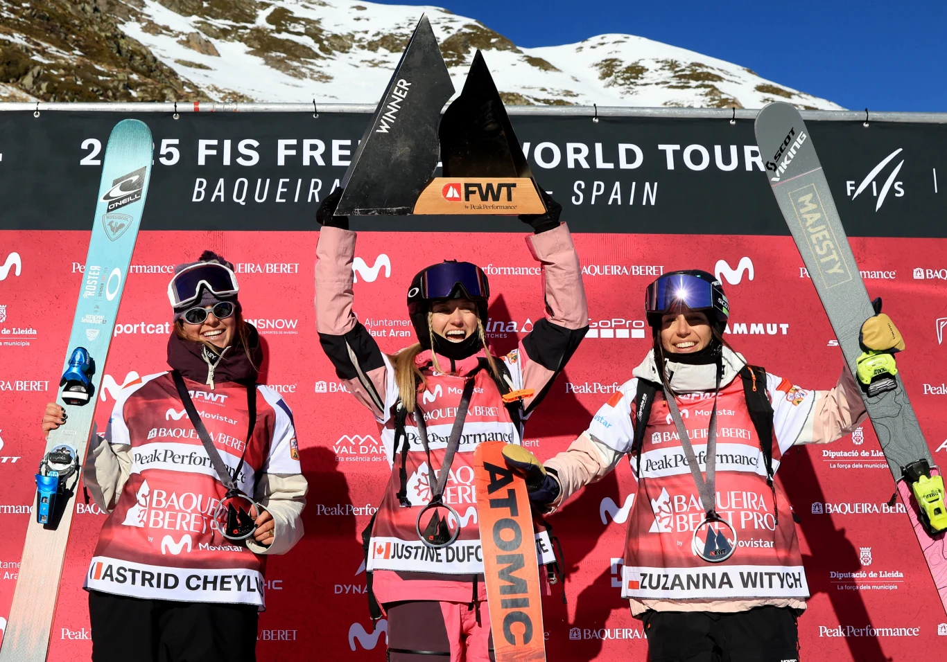 Trzy narciarki stoją na podium zawodów, każda trzyma narty, medal i dyplom, a zwyciężczyni unosi nad głową trofeum. Za nimi znajduje się ścianka z logo sponsorów oraz ośnieżone góry w tle.