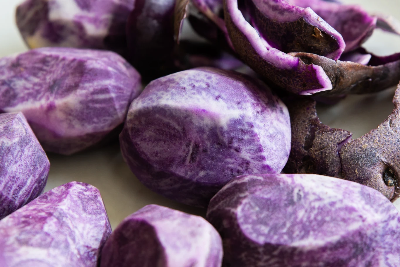 Kilka obranych fioletowych ziemniaków o charakterystycznej marmurkowej strukturze leży na jasnej powierzchni, obok widoczne są fragmenty ich ciemnofioletowej skórki.