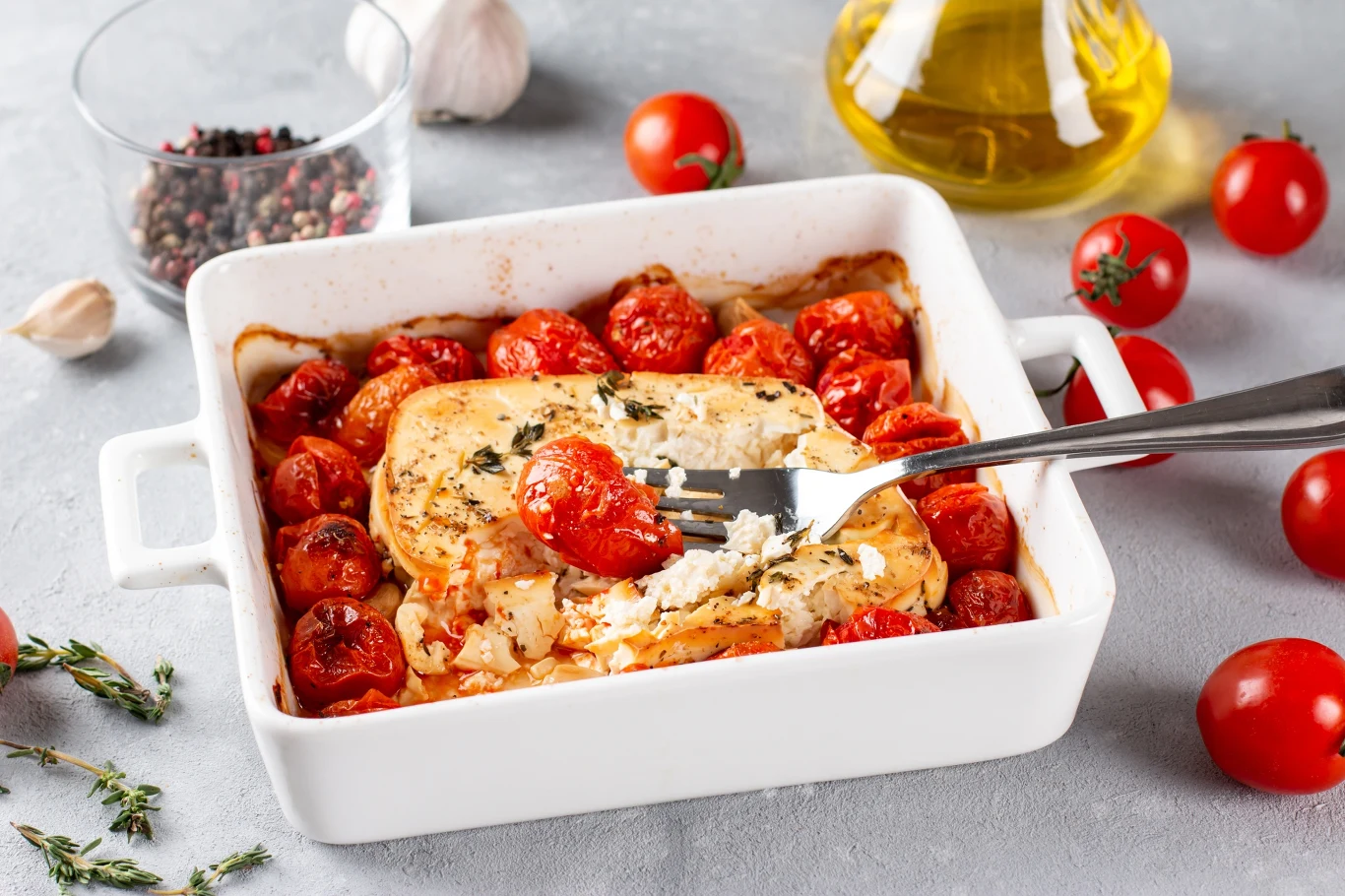 Zapiekany ser feta z pomidorkami koktajlowymi, ziołami i oliwą z oliwek w białym naczyniu żaroodpornym. W tle widoczne świeże składniki: gałązki tymianku, czosnek, oliwa oraz szklanka lekko rozgniecionego pieprzu.