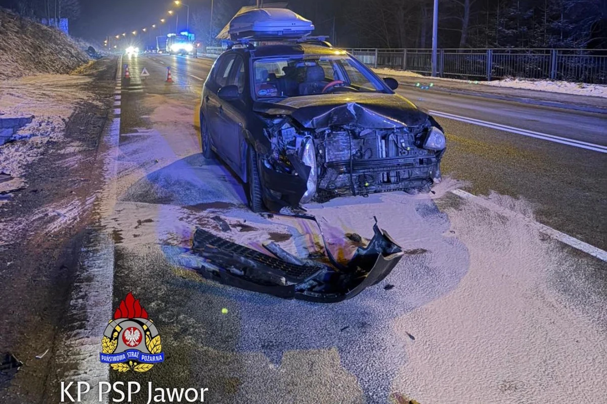 Samochód osobowy z poważnie uszkodzonym przodem stoi na poboczu ośnieżonej drogi w nocy, na jezdni widoczne są ślady rozlanego płynu i rozsypanych materiałów chłonnych, w tle widać inne pojazdy i barierki ochronne.