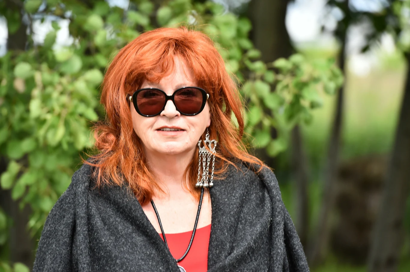 Kobieta o rudych włosach w dużych okularach przeciwsłonecznych, ubrana w ciemny płaszcz i czerwoną bluzkę, stoi na tle zielonych drzew, nosi okazały wisior oraz długie kolczyki.