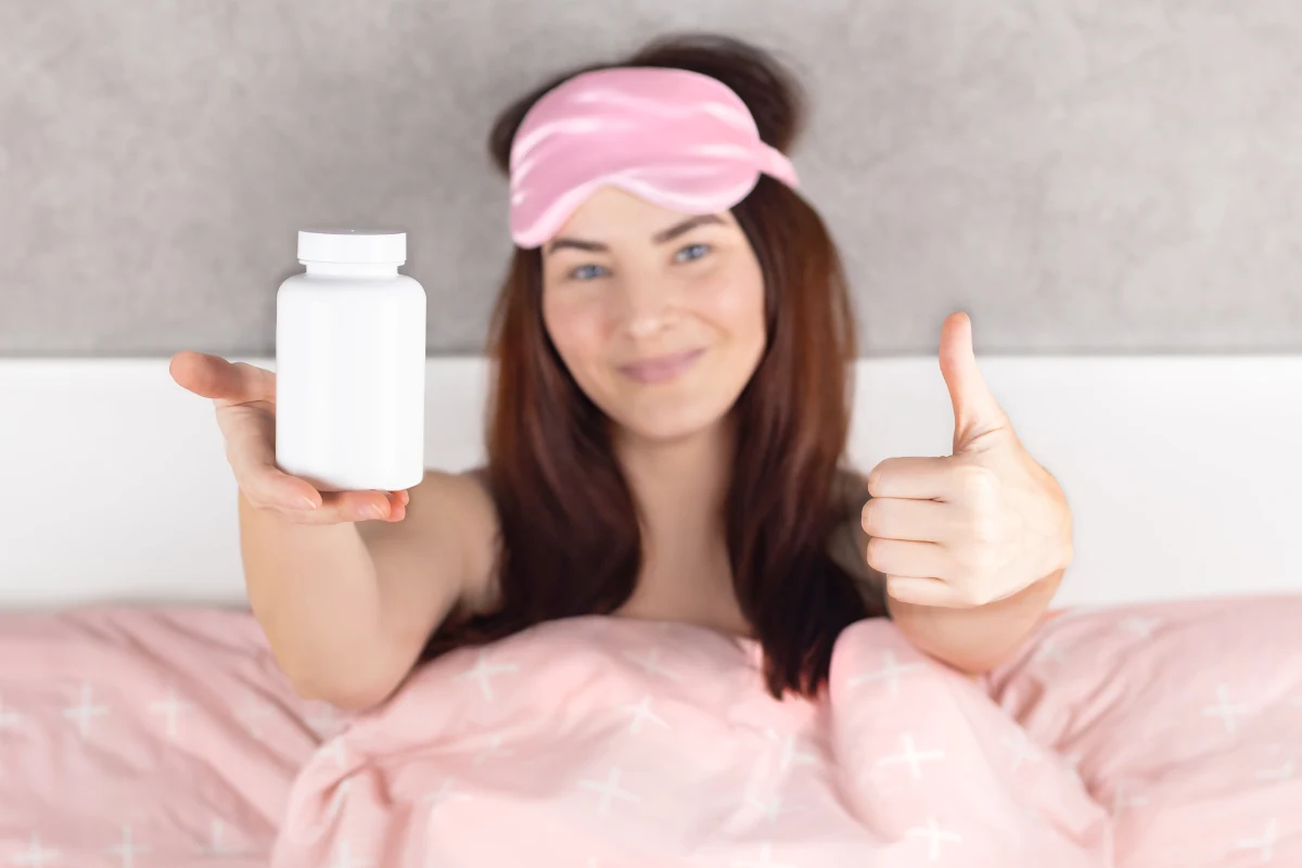Młoda kobieta w piżamie i różowej opasce na oczy siedzi w łóżku, trzymając w jednej dłoni biały pojemnik, najprawdopodobniej na suplementy lub leki, a drugą pokazuje gest okej.