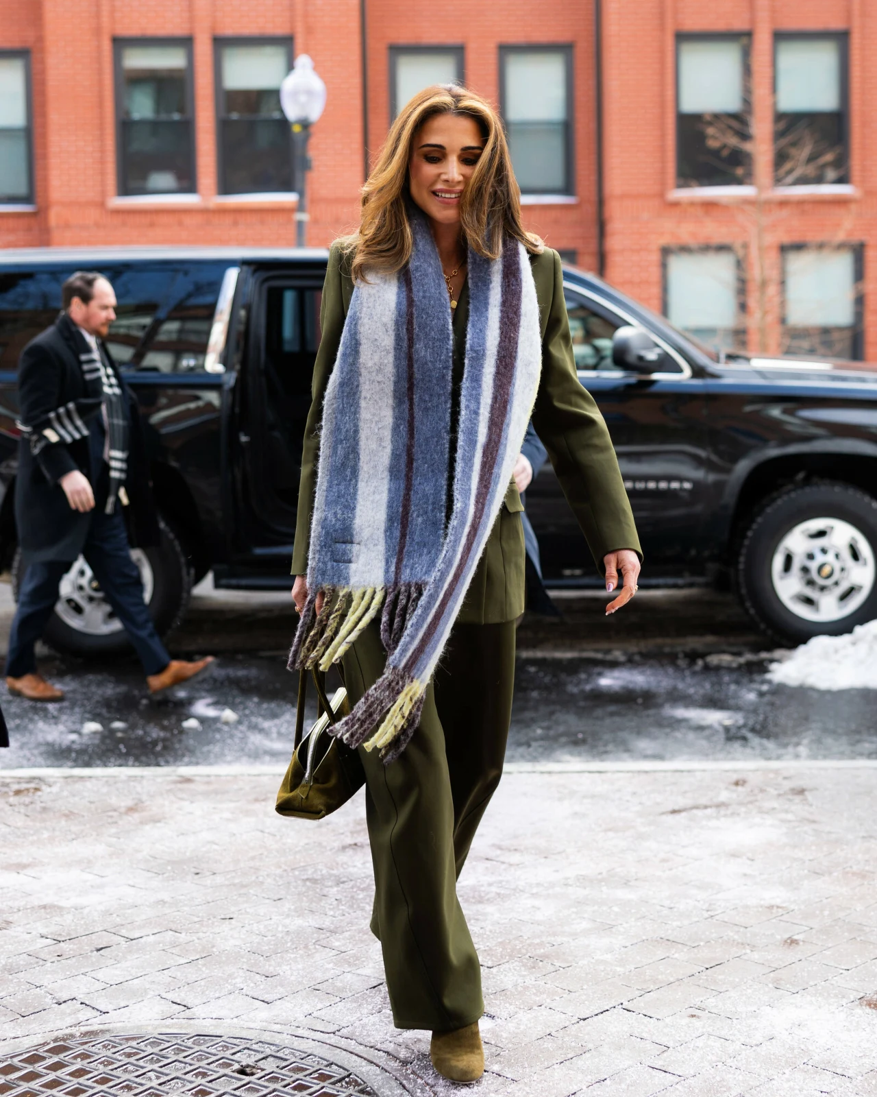 Elegancka kobieta ubrana w zielony garnitur i długi, szeroki szalik w pasy idzie chodnikiem zimową porą, obok zaparkowanego czarnego samochodu terenowego. W tle ceglany budynek z dużymi oknami, leżący śnieg na krawężniku oraz przechodzień.