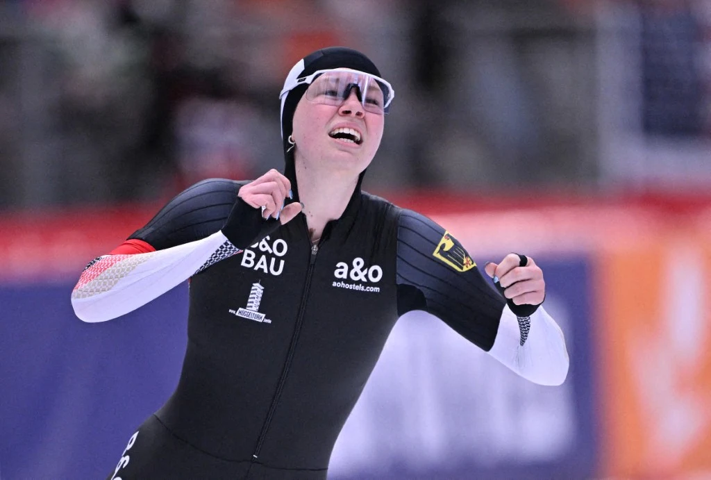 Łyżwiarka szybka w stroju sportowym i goglach unosi ręce w geście triumfu, z wyrazem radości na twarzy, tło rozmyte, wskazujące na obecność publiczności lub innych zawodników.