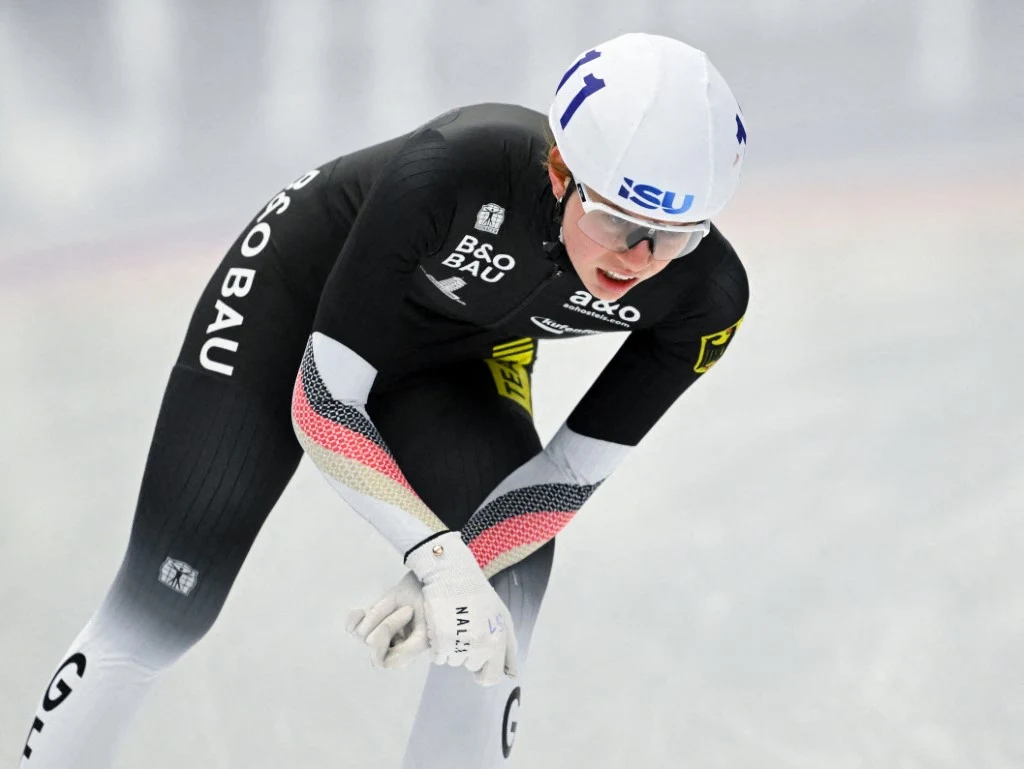 Łyżwiarz szybki w kombinezonie startowym pochylony do przodu podczas jazdy na lodowisku, w białym kasku i okularach ochronnych, skoncentrowany na wyścigu.