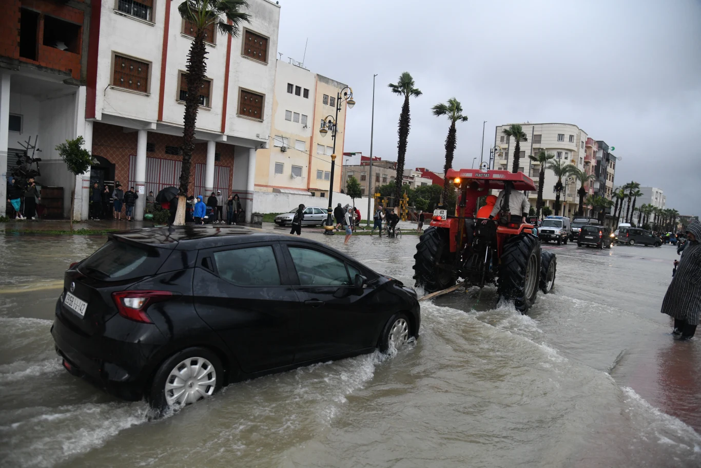 Traktor ciągnie czarny samochód przez zalaną ulicę, w tle widać budynki mieszkalne, palmy oraz przechodniów obserwujących sytuację, deszczowa aura i stojąca woda świadczą o powodzi.