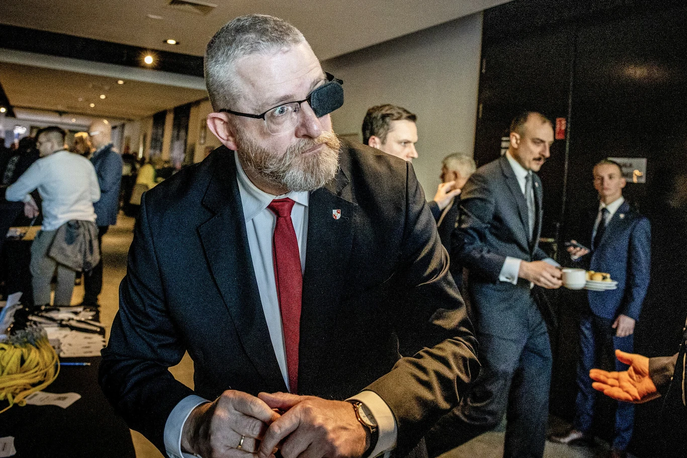 Mężczyzna w garniturze z brodą i przepaską na oku stoi w centrum uwagi podczas formalnego wydarzenia, wokół niego inni mężczyźni w garniturach prowadzą rozmowy lub piją kawę w nowoczesnym wnętrzu.