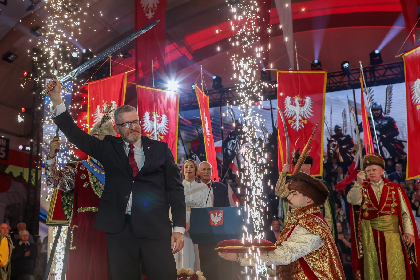 Uroczysta scena z udziałem mężczyzny w garniturze trzymającego w górze miecz, otoczonego przez osoby w historycznych strojach i symbole narodowe Polski, w tle biało-czerwone flagi z orłem oraz efekty świetlne i iskry podkreślające podniosłość wydarzenia.