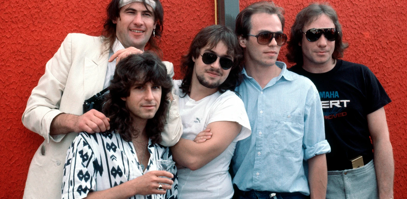 Marillion w 1980 roku Pięciu mężczyzn pozuje razem na tle czerwonej ściany, część z nich nosi okulary przeciwsłoneczne, a jeden ubrany jest w jasny garnitur i opaskę na głowie, wyglądają na członków zespołu muzycznego lub grupy przyjaciół.