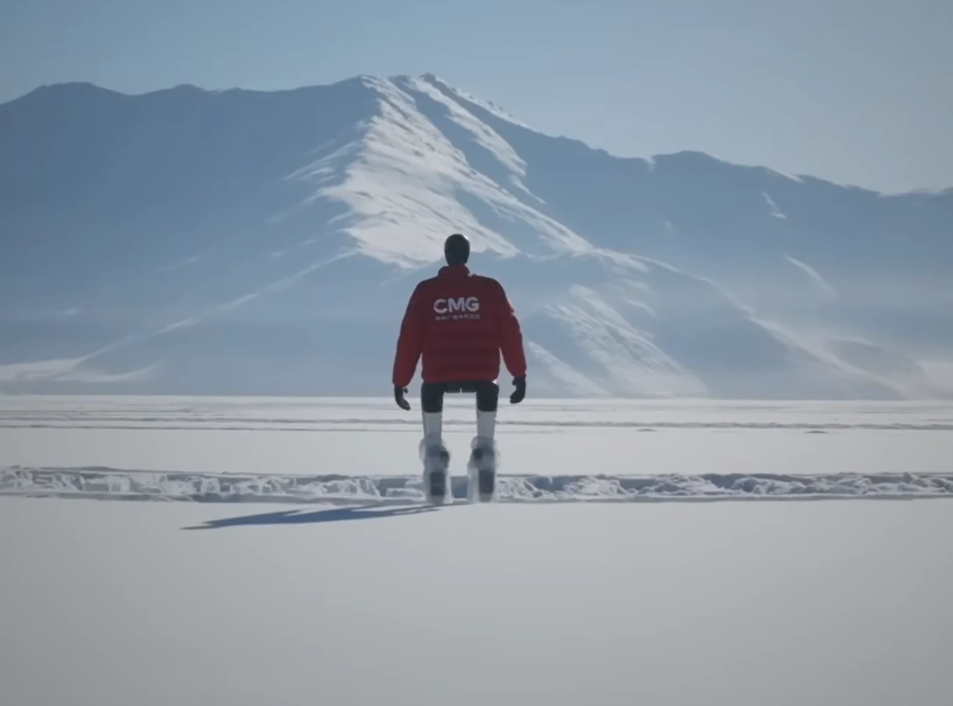 Postać w czerwonej kurtce z napisem CMG stoi na rozległej, ośnieżonej równinie, w tle wysokie, ośnieżone góry wyłaniające się na horyzoncie pod jasno-niebieskim niebem.