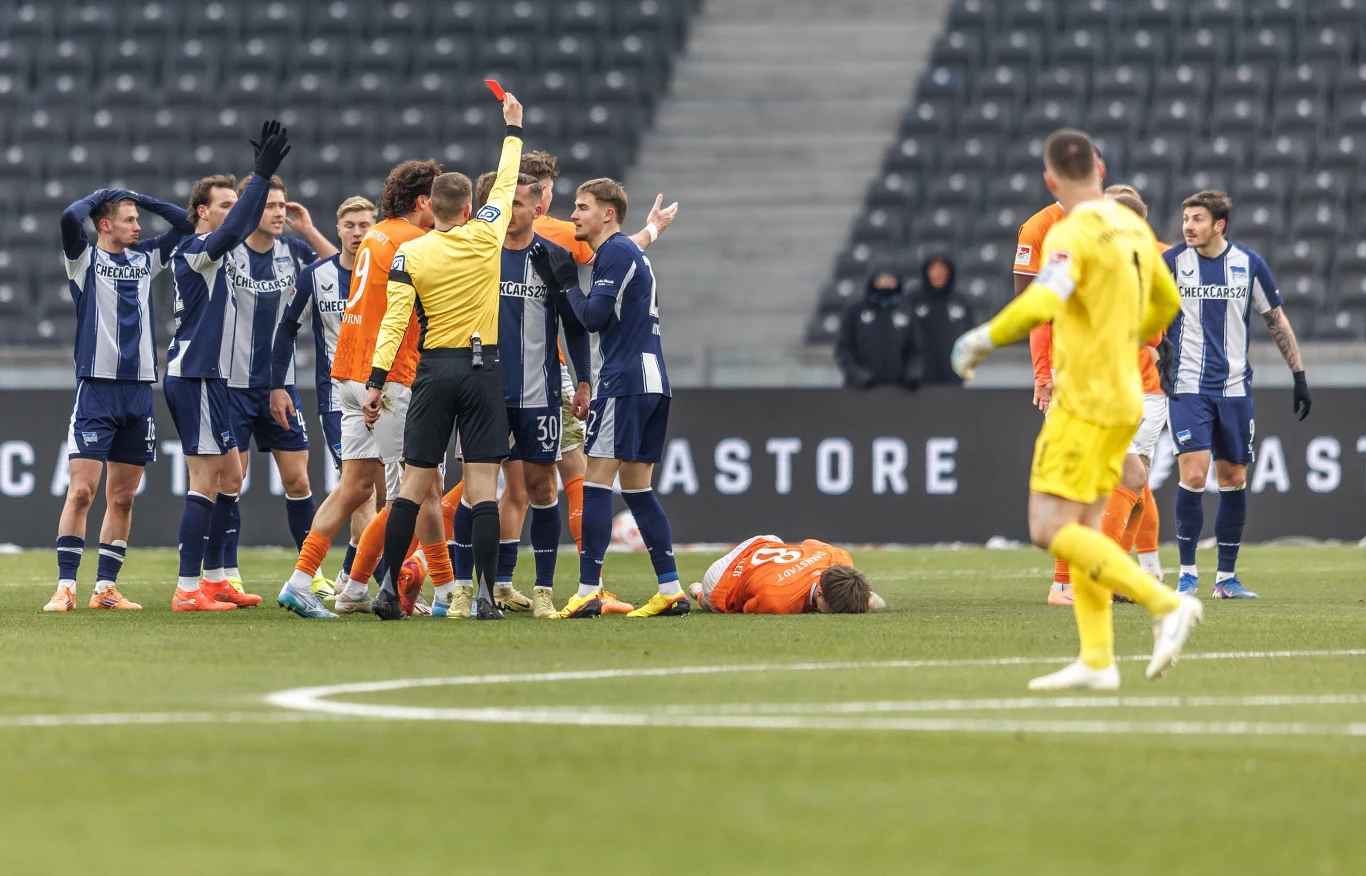 Sędzia podnosi czerwoną kartkę w kierunku piłkarza w pomarańczowym stroju, który leży na murawie, otoczony przez innych zawodników w ciemnoniebieskich oraz pomarańczowych strojach. W tle widoczny pusty sektor trybun.