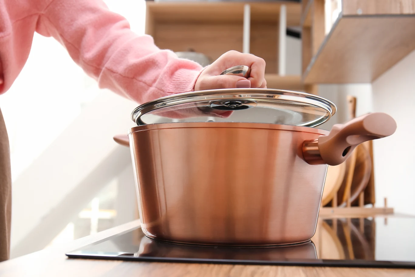 Nowoczesne garnki miedziane są bezpieczne dla zdrowia pod warunkiem, że prawidłowo się z nich korzysta Kuchenna scena z osobą w różowym swetrze podnoszącą pokrywkę miedzianego garnka stojącego na indukcyjnej płycie grzewczej. W tle drewniane elementy i nowoczesna kuchnia.