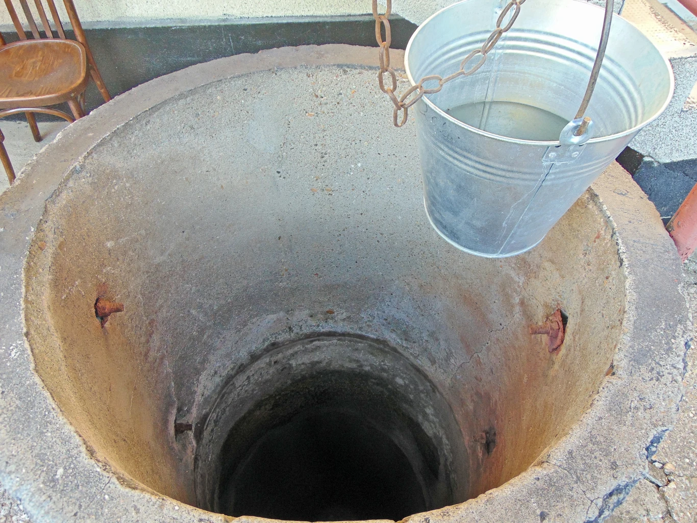 Otwarta studnia z betonowym obramowaniem, do której spuszczone jest metalowe wiadro na łańcuchu. W tle widoczne stare krzesło oraz fragment ściany.