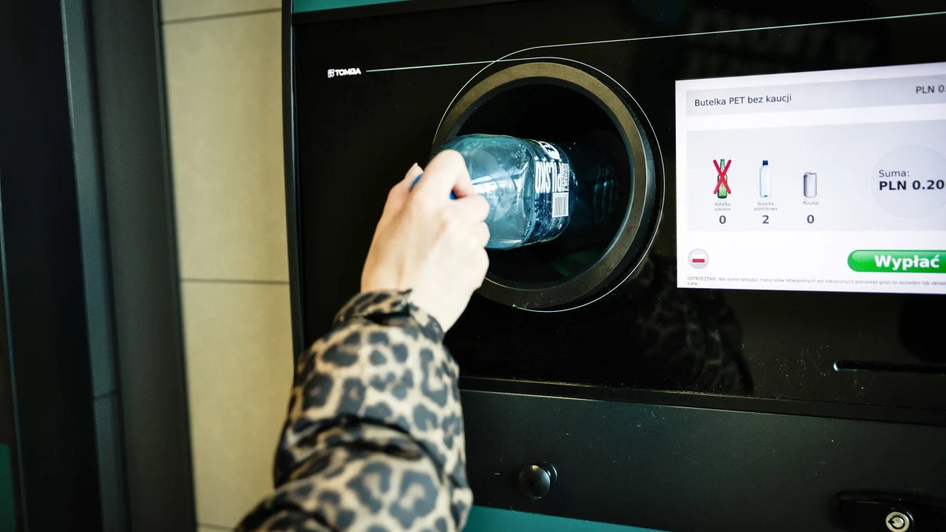 Ręka w kurtce we wzór panterki wkłada plastikową butelkę po wodzie mineralnej do automatu przyjmującego opakowania zwrotne, po prawej stronie widoczny jest ekran wyświetlający informacje o liczbie oddanych butelek.