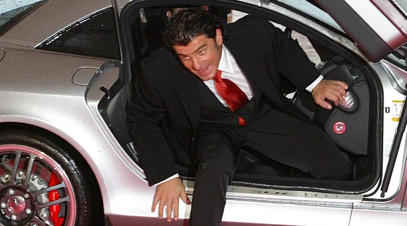 Elegancko ubrany mężczyzna w garniturze i czerwonym krawacie wychodzi pochylony z luksusowego srebrnego samochodu sportowego z widocznymi czerwonymi felgami.