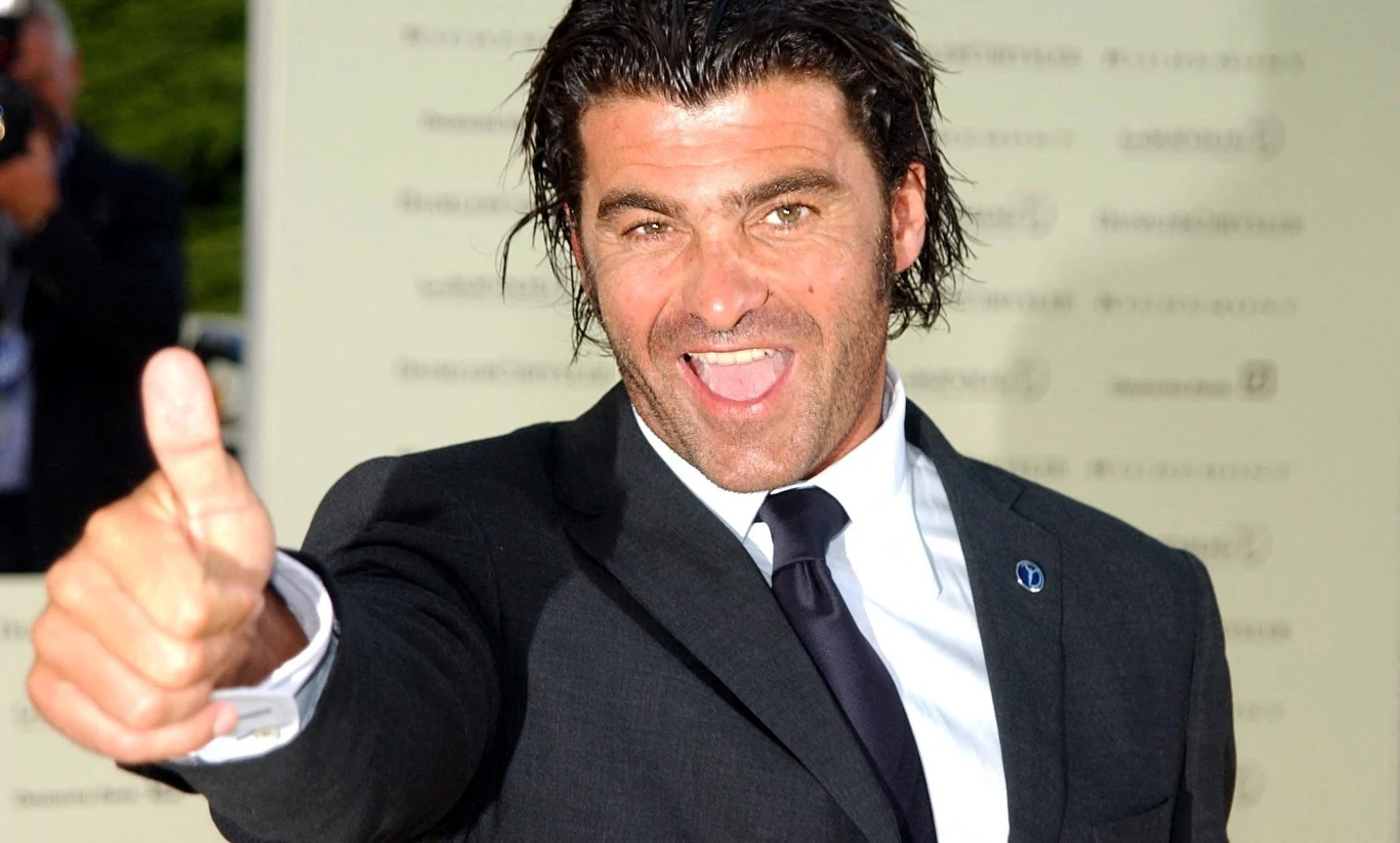 Mężczyzna w ciemnym garniturze z białą koszulą i krawatem pozuje z szerokim uśmiechem, pokazując uniesiony kciuk w geście aprobaty.