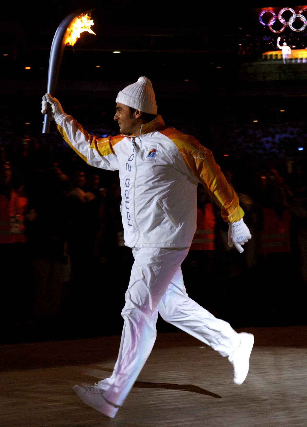 Mężczyzna ubrany w biały strój sportowy z żółtymi akcentami biegnie z zapaloną olimpijską pochodnią na arenie podczas ceremonii, w tle widownia i symboliczne pierścienie olimpijskie.