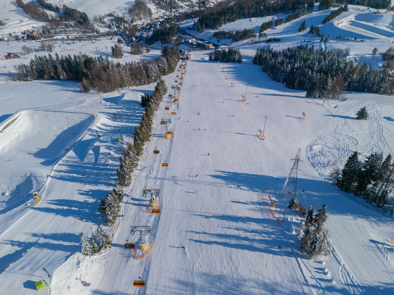 Stok narciarski widziany z lotu ptaka, otoczony gęstym lasem i otwartymi przestrzeniami, z widocznymi wyciągami narciarskimi oraz kilkoma narciarzami korzystającymi z trasy. W tle pagórkowaty, zimowy krajobraz z fragmentami zabudowań.