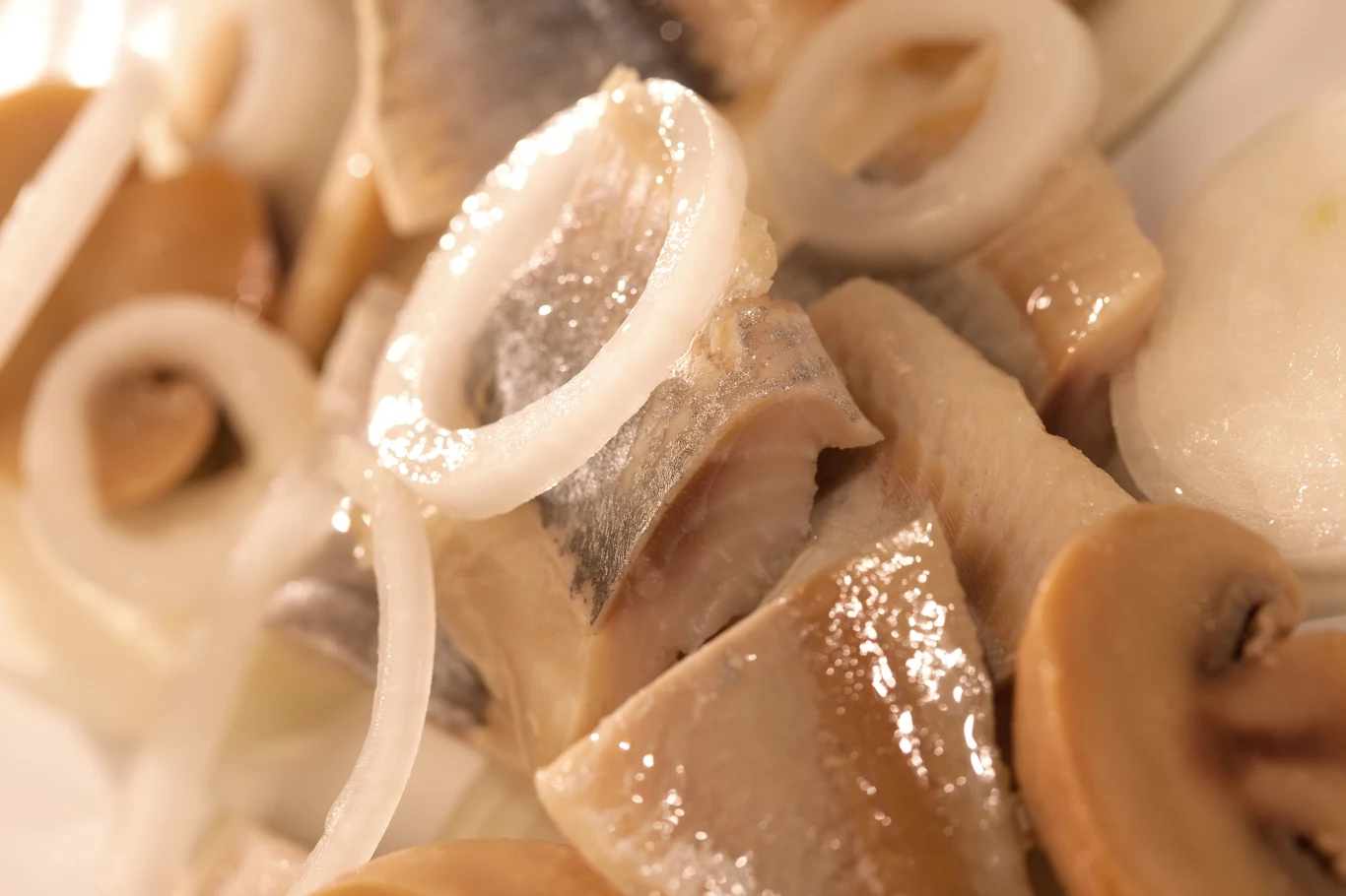 Kawałki śledzia z błyszczącą skórą ułożone obok plasterków cebuli i kawałków marynowanych pieczarek.