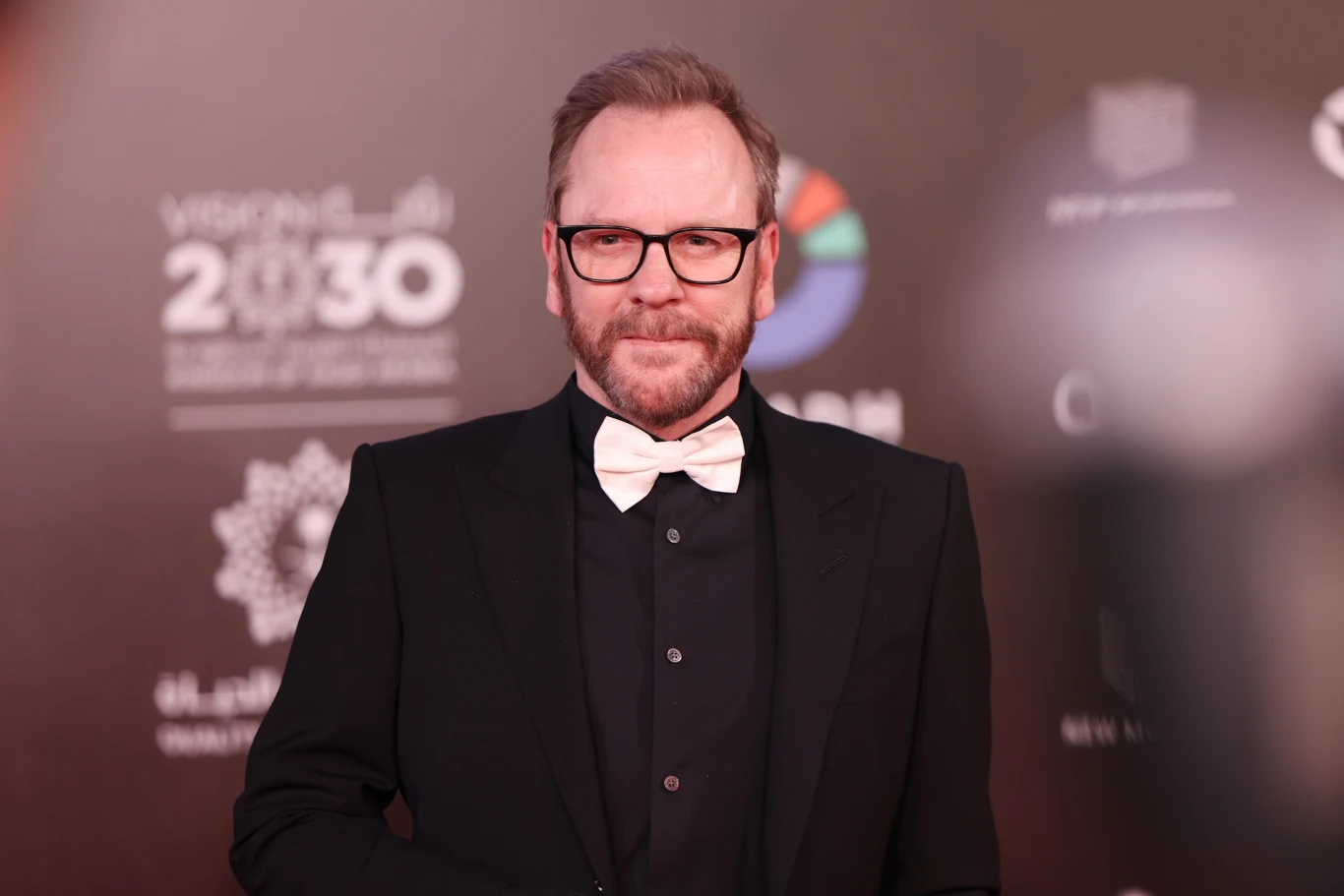 Kiefer Sutherland przyjedzie do Warszawy Mężczyzna w okularach, ubrany w czarny garnitur i białą muchę, stoi na tle ściany z logotypami oraz napisem Vision 2030.
