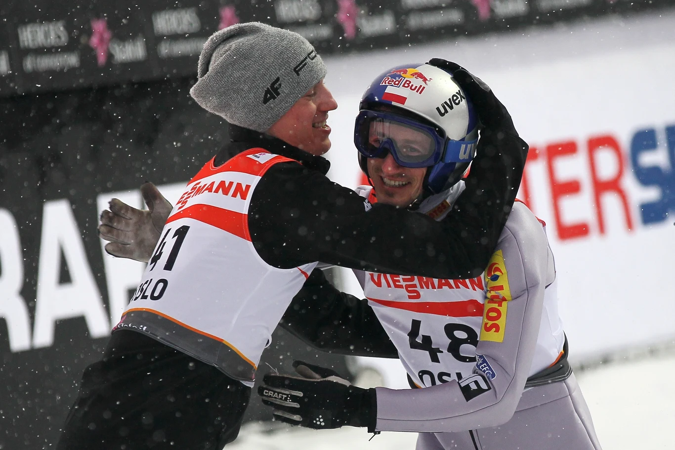 Dwóch skoczków narciarskich w kombinezonach i kaskach, przy śnieżnej scenerii, ściska się w geście radości i gratulacji po zawodach. W tle widoczne są bannery sponsorskie.