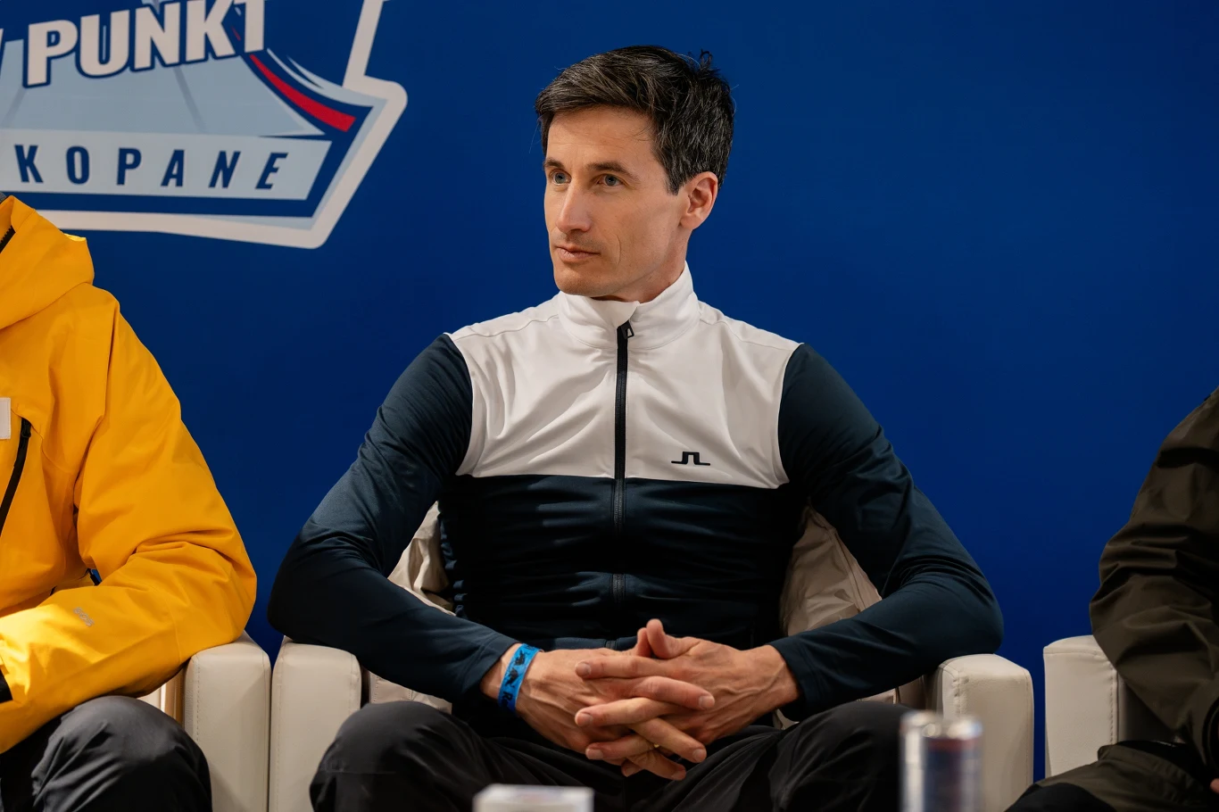 Mężczyzna w sportowym stroju siedzący na białym fotelu, z założonymi dłońmi na kolanach. W tle widoczne niebieskie tło z logo i napisem Zakopane, po bokach fragmenty innych postaci.