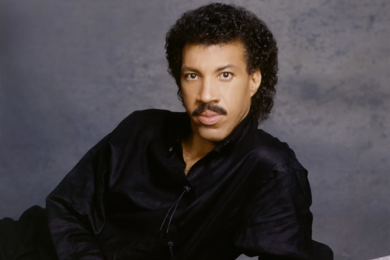 Lionel Richie w latach 70. zasłynął balladą "Easy" Mężczyzna z bujnymi kręconymi włosami i wąsem ubrany w czarną koszulę, patrzący wprost w obiektyw, pozujący na tle neutralnej, szarej scenerii.