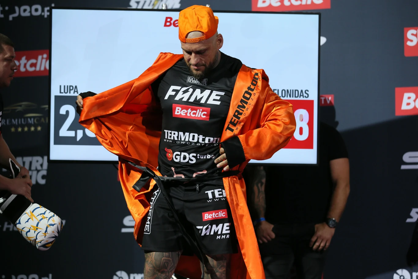 Mężczyzna w sportowym stroju promocyjnym oraz pomarańczowym szlafroku i czapce z daszkiem tyłem do przodu stoi na tle tablicy z logo sponsorów podczas oficjalnego ważenia przed galą sportową.