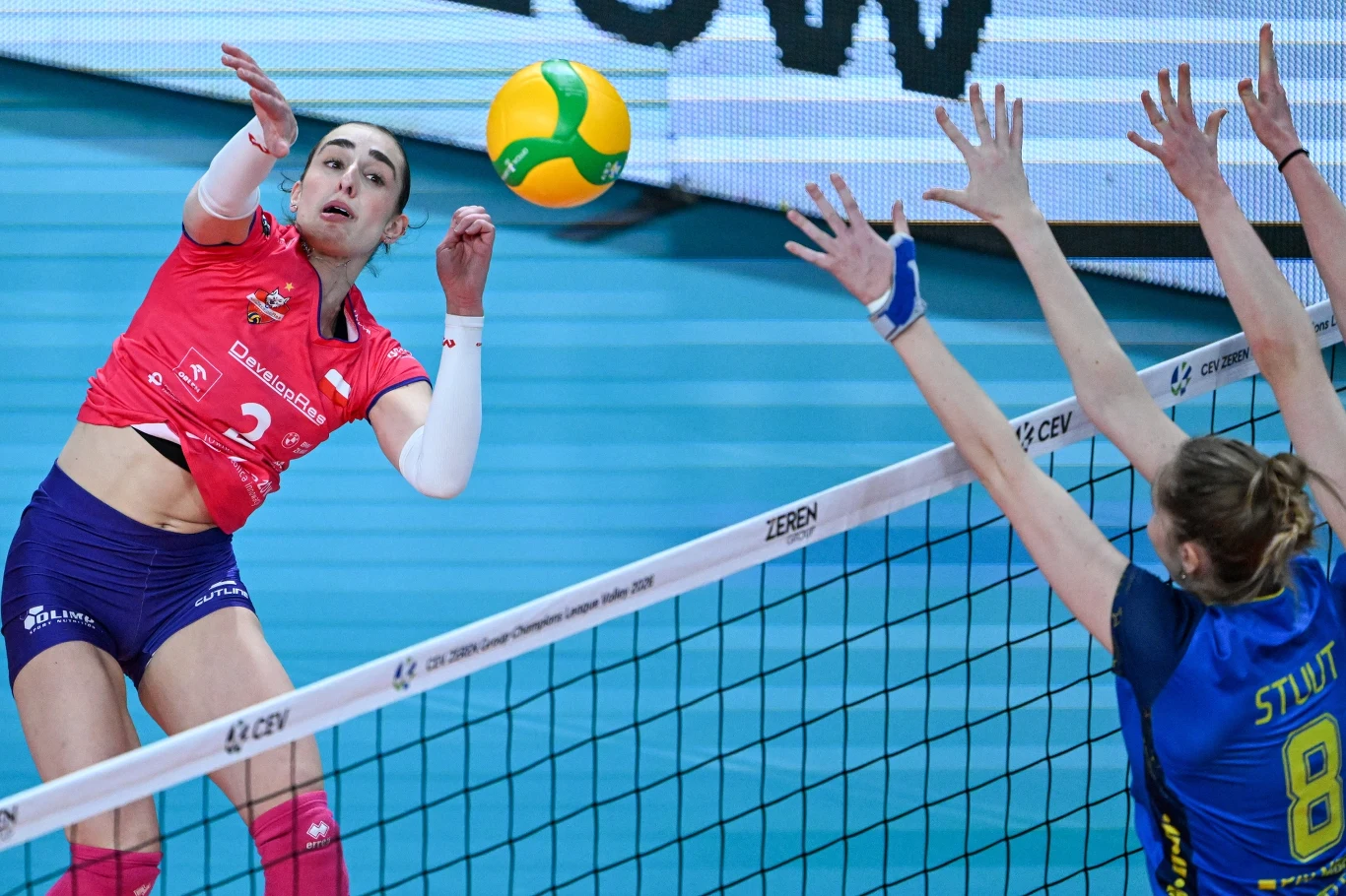 Zawodniczka w różowej koszulce wykonuje atak podczas meczu siatkówki, a po drugiej stronie siatki dwie rywalki w niebieskich strojach przygotowują się do bloku. Piłka znajduje się tuż nad siatką, a sportowcy są skoncentrowani na akcji.