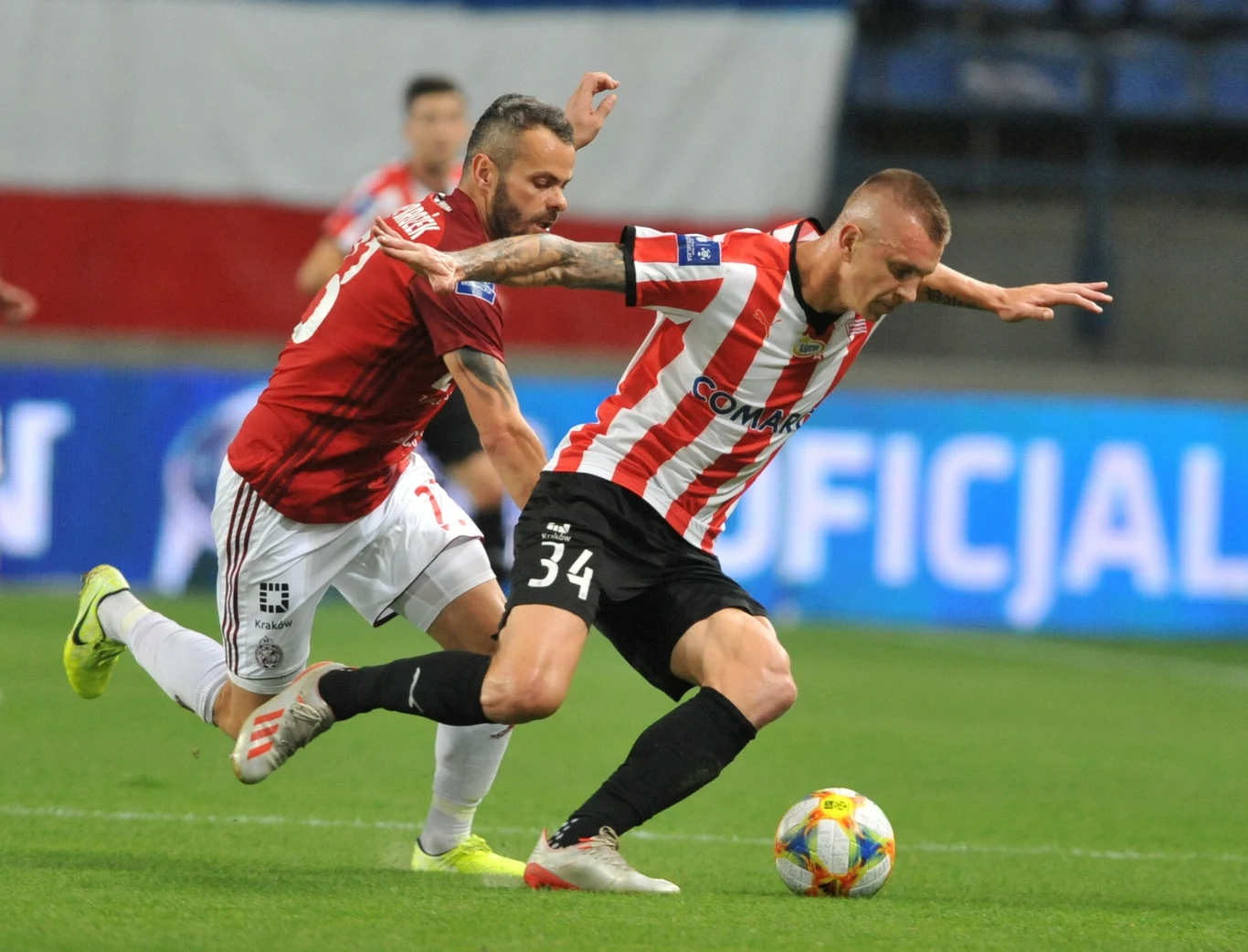 Dwóch piłkarzy walczy o piłkę na murawie, jeden z zawodników w czerwono-białej koszulce próbuje utrzymać się na nogach, podczas gdy drugi w czerwonej koszulce stara się go zatrzymać. W tle widoczna flaga i rozmyci widzowie.