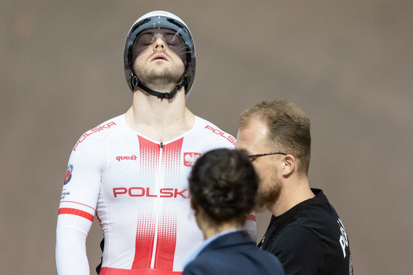 Mateusz Rudyk (zdj. archiwalne) Kolarz torowy w biało-czerwonym stroju z napisem Polska przygotowuje się do startu, stoi z zamkniętymi oczami i skupieniem na twarzy, obok niego dwie osoby z obsługi technicznej.