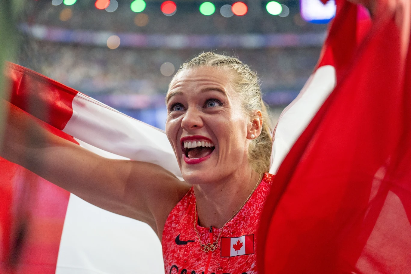 Szczęśliwa sportsmenka w stroju reprezentacji Kanady trzymająca dużą kanadyjską flagę, z szerokim uśmiechem świętuje sukces na stadionie w otoczeniu jasnych świateł i rozmytych widzów.