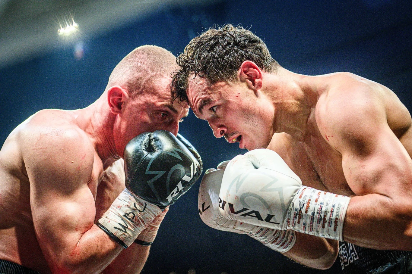 Dwóch bokserów w trakcie zaciętej walki, skupionych na sobie, z rękawicami uniesionymi tuż przy twarzach. Intensywne spojrzenia i napięte mięśnie podkreślają emocje i rywalizację.