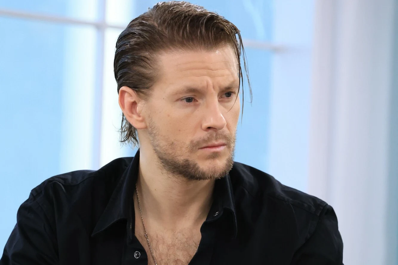 Mężczyzna o jasnej cerze i ciemnych włosach zaczesanych do tyłu, ubrany w ciemną koszulę, siedzi w jasnym pomieszczeniu z dużym oknem w tle, patrzy lekko w bok z poważnym wyrazem twarzy.