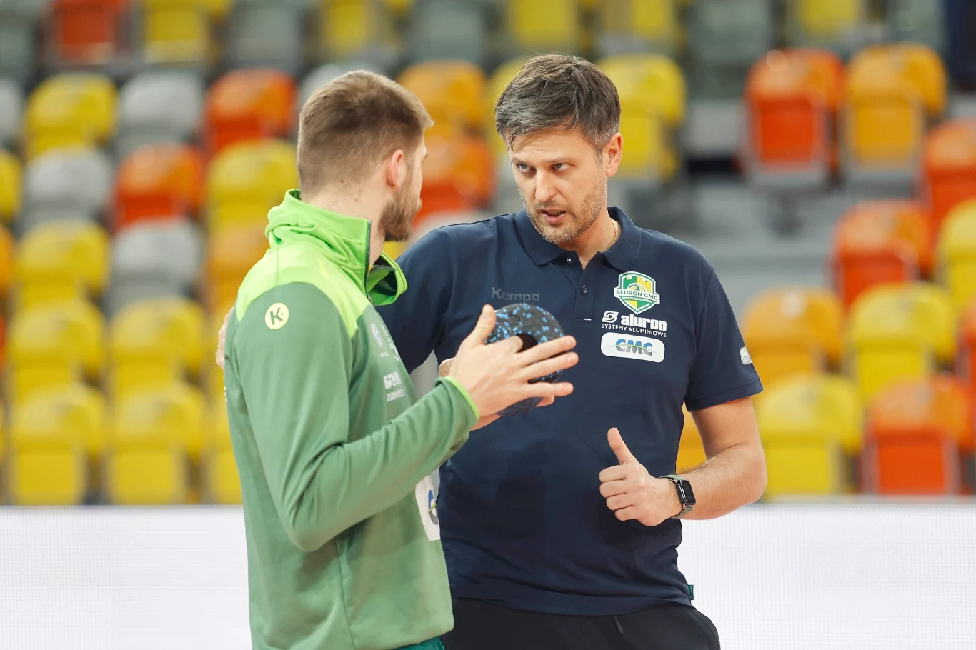 Dwóch mężczyzn w sportowych ubraniach rozmawia na hali sportowej, jeden z nich trzyma piłkę ręczną, drugi gestykuluje kciukiem w górę, w tle widoczne kolorowe siedzenia trybun.