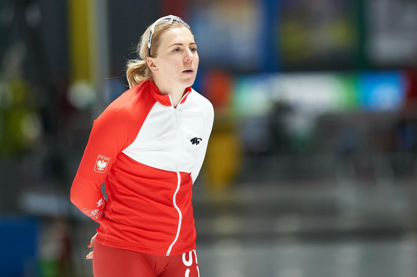 Natalia Czerwonka Łyżwiarka w czerwono-białym stroju reprezentacyjnym z godłem Polski na piersi, w tle rozmyte wnętrze hali sportowej.