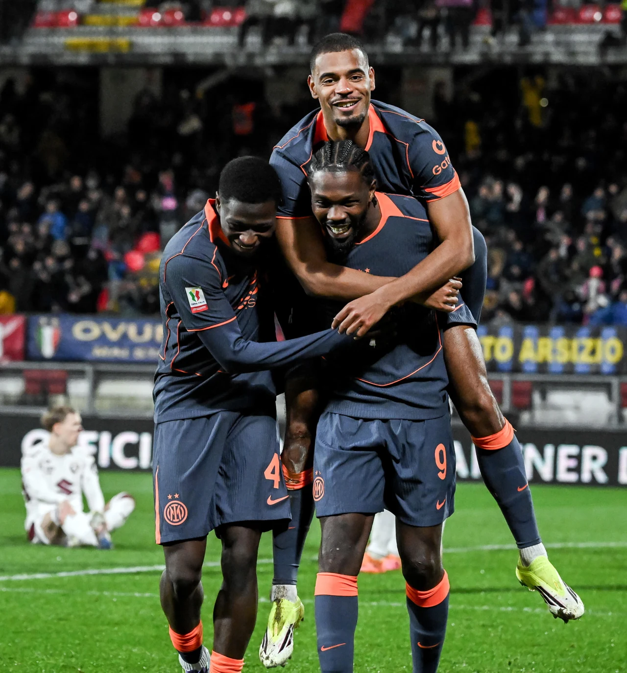 Trzech piłkarzy w granatowych strojach z pomarańczowymi elementami świętuje na murawie stadionu, dwóch z nich stoi, a trzeci w radosnym geście wskakuje na plecy kolegi, w tle rozmyta publiczność i fragmenty trybun.