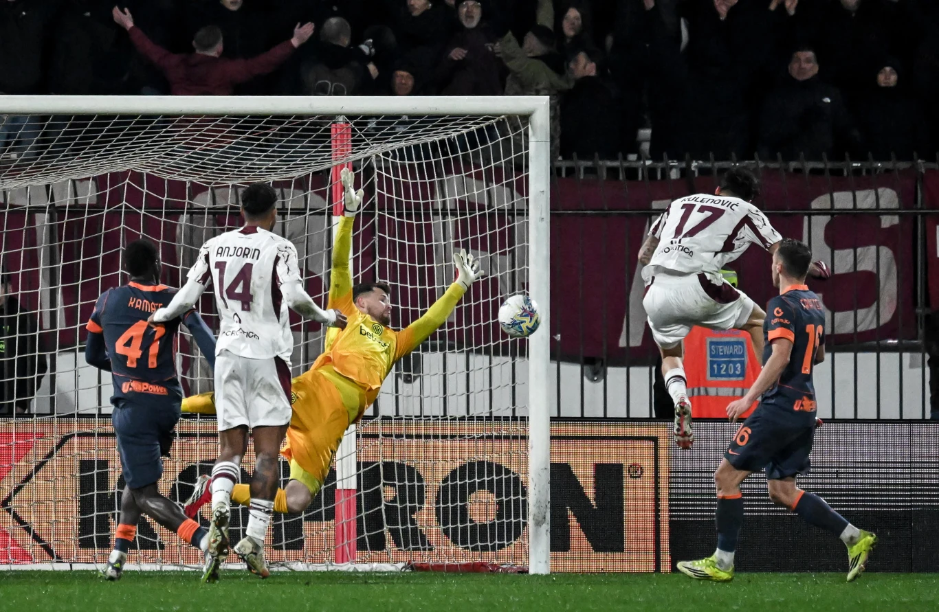 Piłkarz w białym stroju wyskakuje do piłki w polu karnym, próbując zdobyć bramkę, podczas gdy bramkarz w żółtym stroju rzuca się, by obronić strzał. W tle kilku innych zawodników oraz kibice na trybunach.