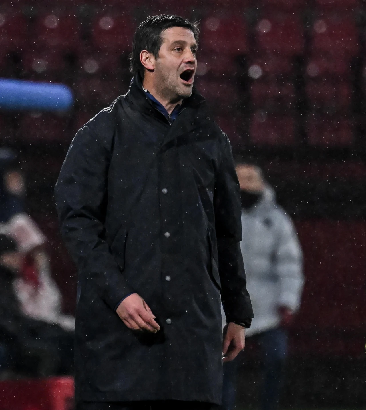 Mężczyzna w ciemnym płaszczu krzyczy lub wydaje polecenia, stojąc na tle rozmytego stadionu. W tle widoczne są niewyraźne postacie oraz rzędy czerwonych krzeseł.