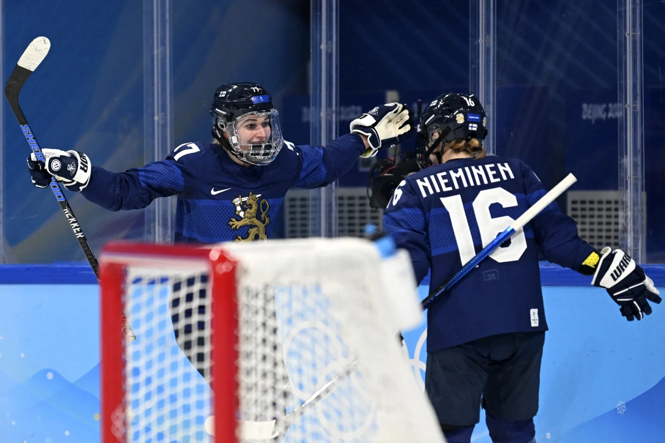 Problemy hokeistek z Finlandii. Wirus w kadrze Dwóch zawodników fińskiej drużyny hokeja na lodzie w niebieskich strojach świętuje zdobycie bramki przed bramką przeciwnika, jeden z nich ma uniesione ręce w geście triumfu, widoczne nazwiska zawodników na koszulkach oraz kij hokejowy.