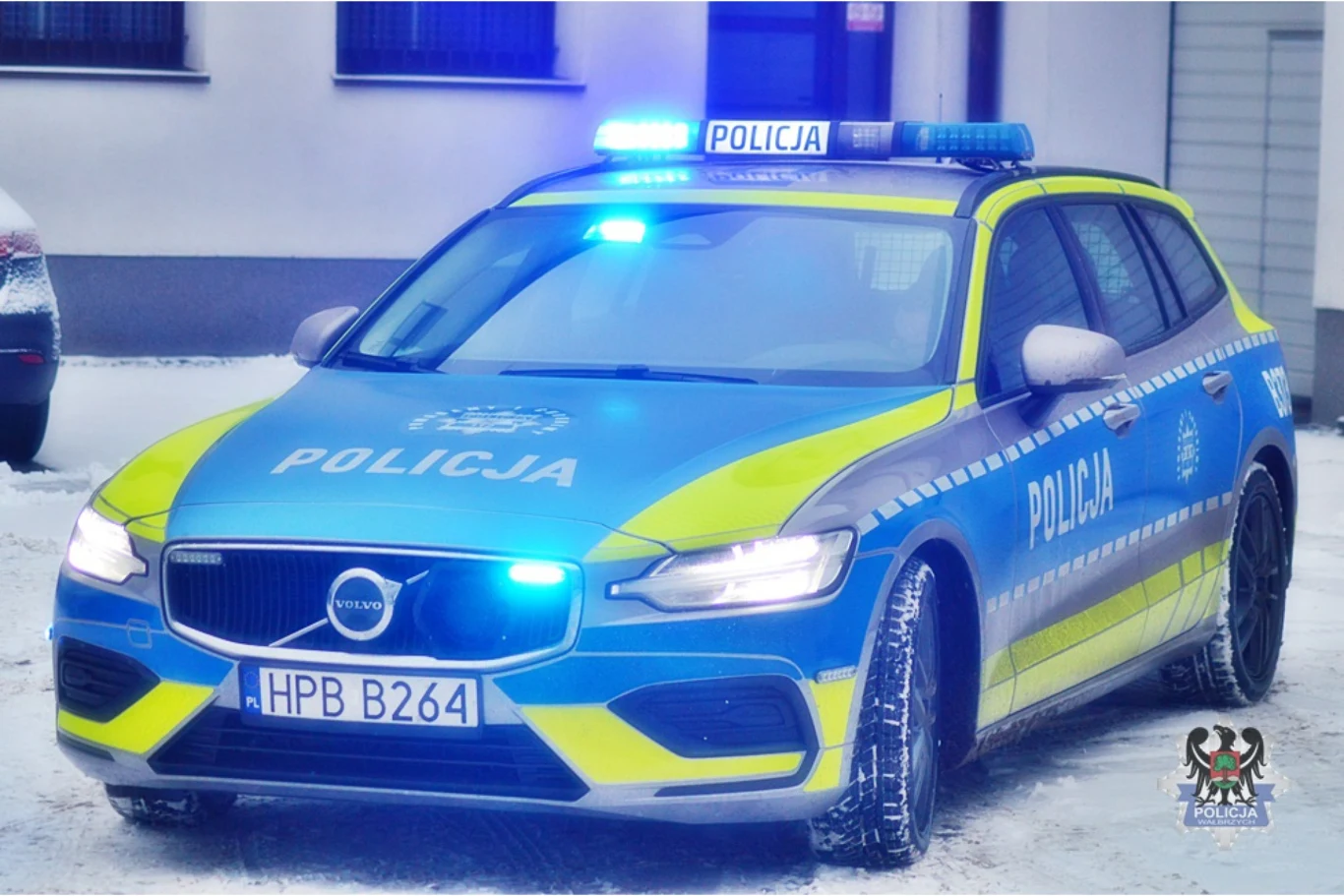 Policyjny radiowóz marki Volvo z włączonymi niebieskimi światłami ostrzegawczymi, oznakowany napisem 'POLICJA', stojący na ośnieżonej ulicy przed budynkiem. W prawym dolnym rogu widoczny herb.