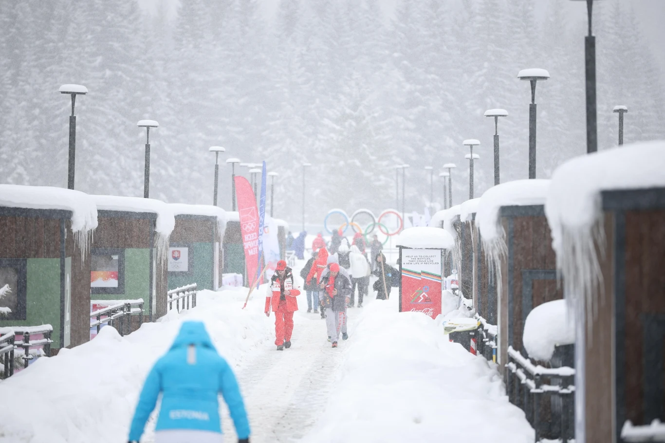 Wioska olimpijska Grupa osób ubranych w zimowe stroje porusza się ośnieżoną alejką pomiędzy drewnianymi budynkami, w tle widoczne olimpijskie koła oraz gęsty las pokryty śniegiem.