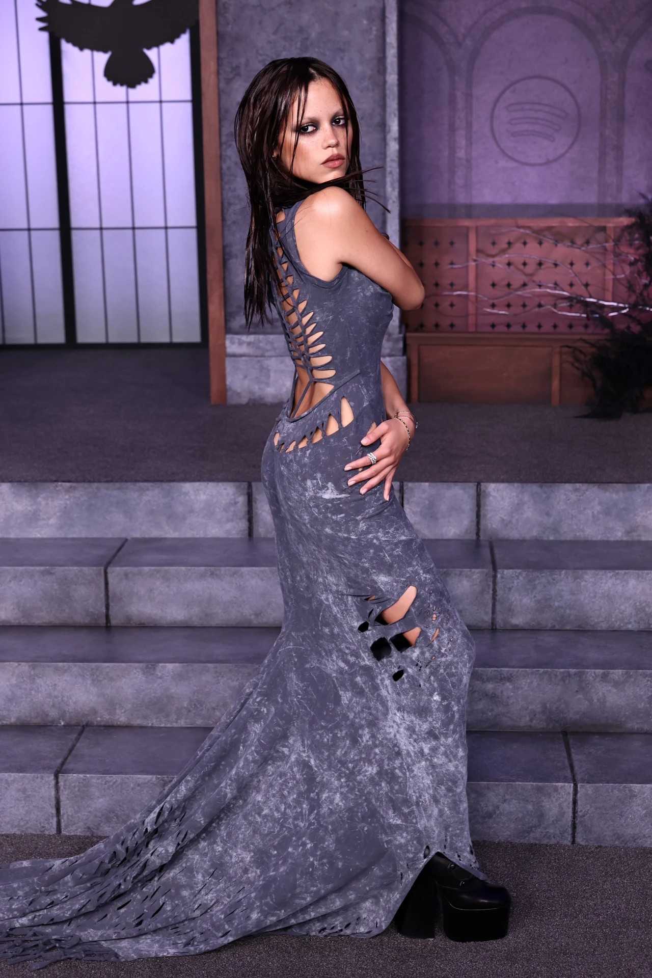 Młoda kobieta w długiej, szarej sukni z licznymi wycięciami i rozdarciami stoi bokiem na schodach we wnętrzu inspirowanym gotykiem, z mrocznym makijażem oraz mokrymi włosami, na tle szarych murów i stylizowanych okien.