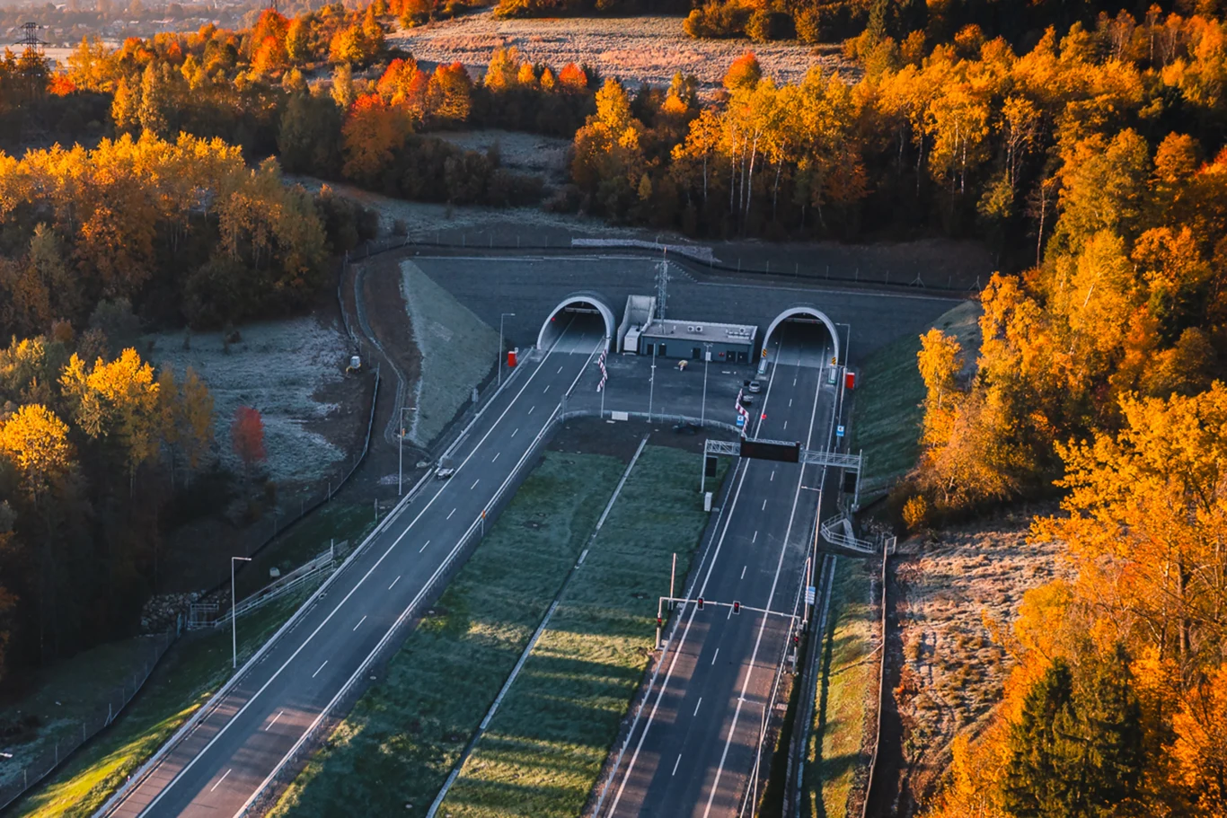 Droga ekspresowa prowadząca przez tunel otoczona jesiennym, kolorowym lasem, podkreślona kontrastem między zielenią trawy a żółtymi i pomarańczowymi liśćmi drzew.
