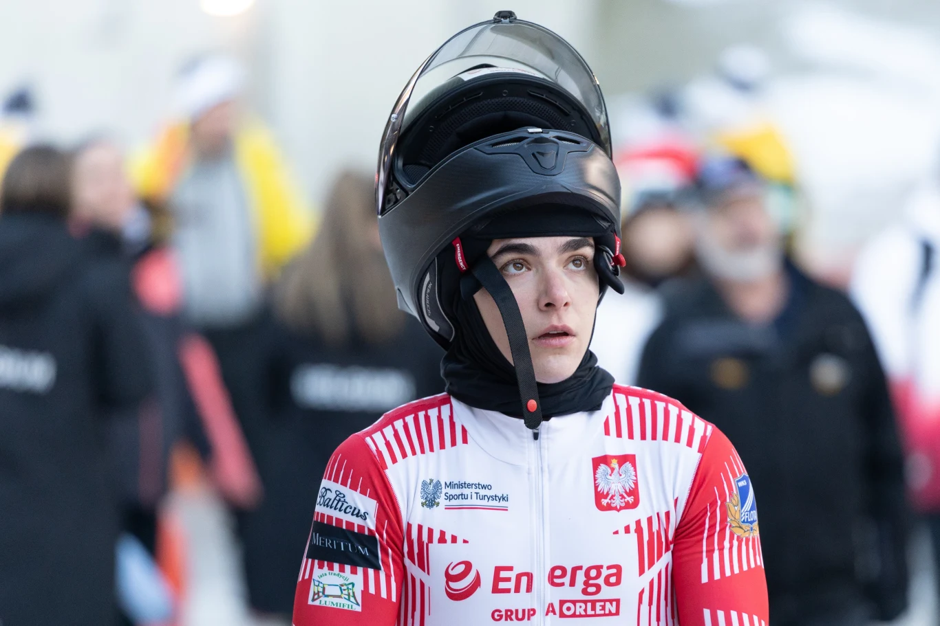Zawodnik w kombinezonie sportowym z logo reprezentacji Polski i sponsorów, trzymający kask luge na głowie, skoncentrowany, na tle rozmytej grupy osób w zimowym otoczeniu.