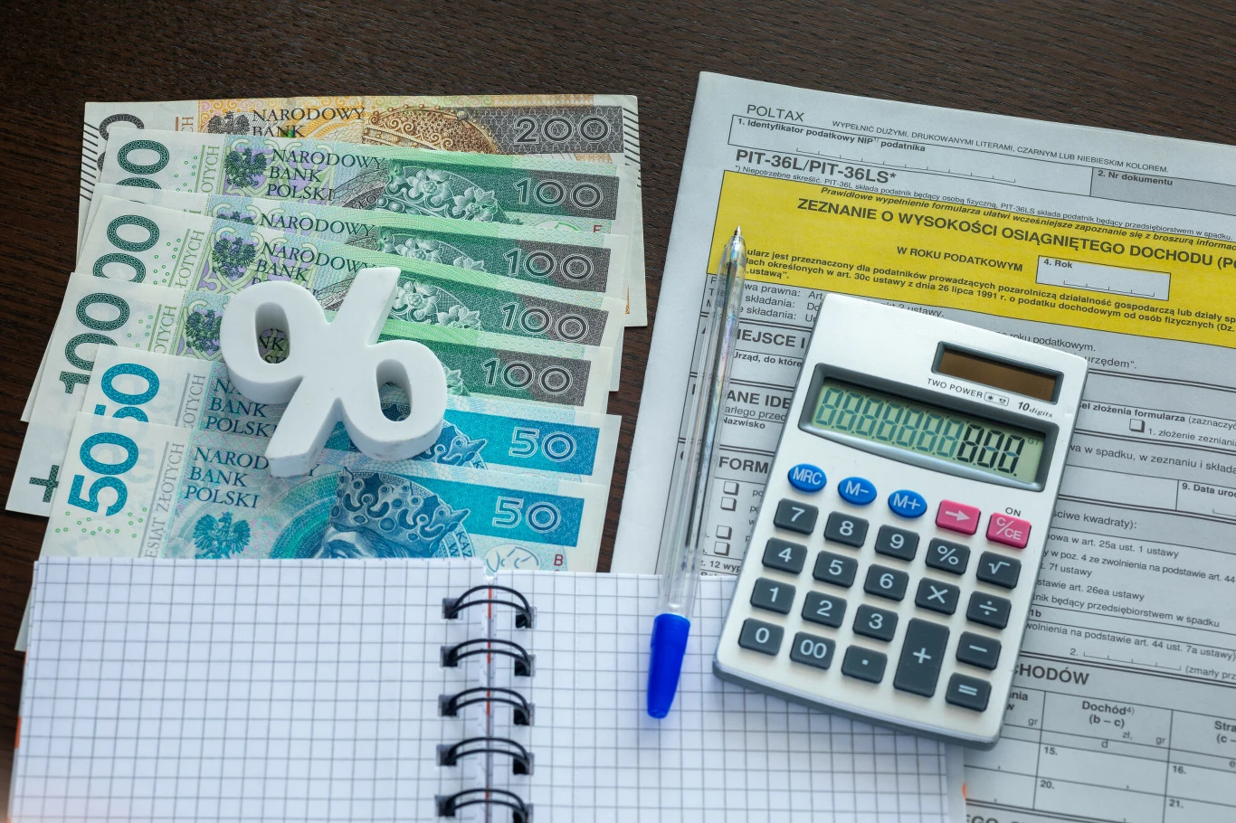 Zeznanie roczne należy złożyć najpóźniej do 30 kwietnia 2026 roku. Banknoty polskie, formularze podatkowe, długopis, kalkulator, notes w kratkę oraz symbol procentu tworzą zestawienie nawiązujące do tematyki podatków i rozliczeń finansowych.