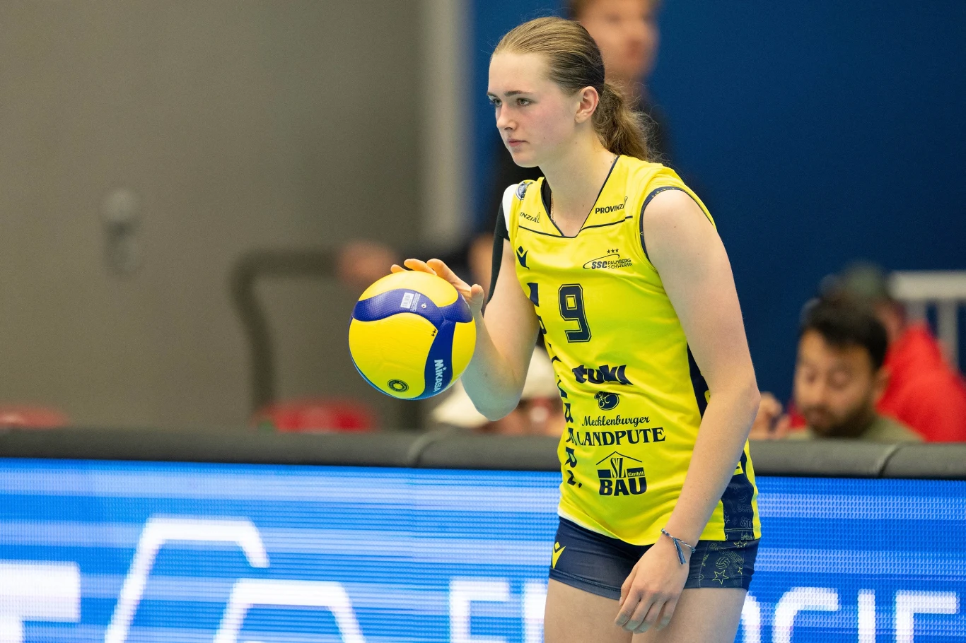 Siatkarka w żółtym stroju sportowym z numerem 9 stoi na boisku, trzymając piłkę do siatkówki w prawej ręce. W tle widać nieostrych kibiców oraz elementy hali sportowej.