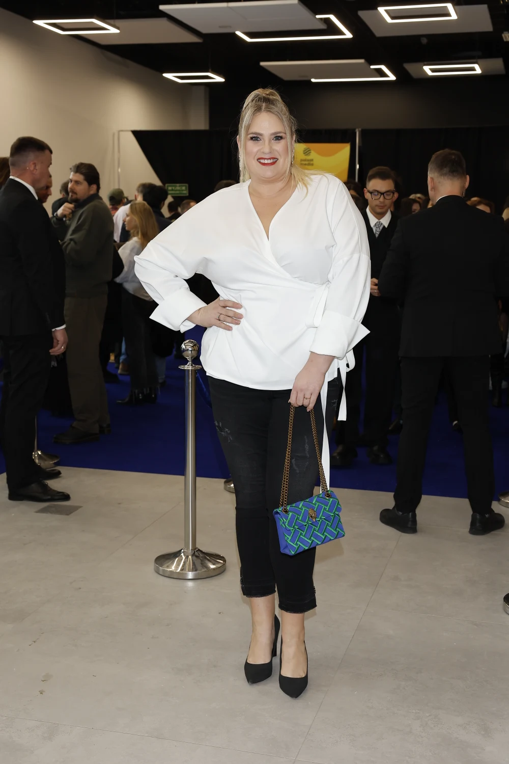 Kobieta w białej koszuli i czarnych spodniach stoi na pierwszym planie, trzymając torebkę o niebiesko-zielonym wzorze, w tle liczna grupa ludzi podczas uroczystego wydarzenia lub konferencji.