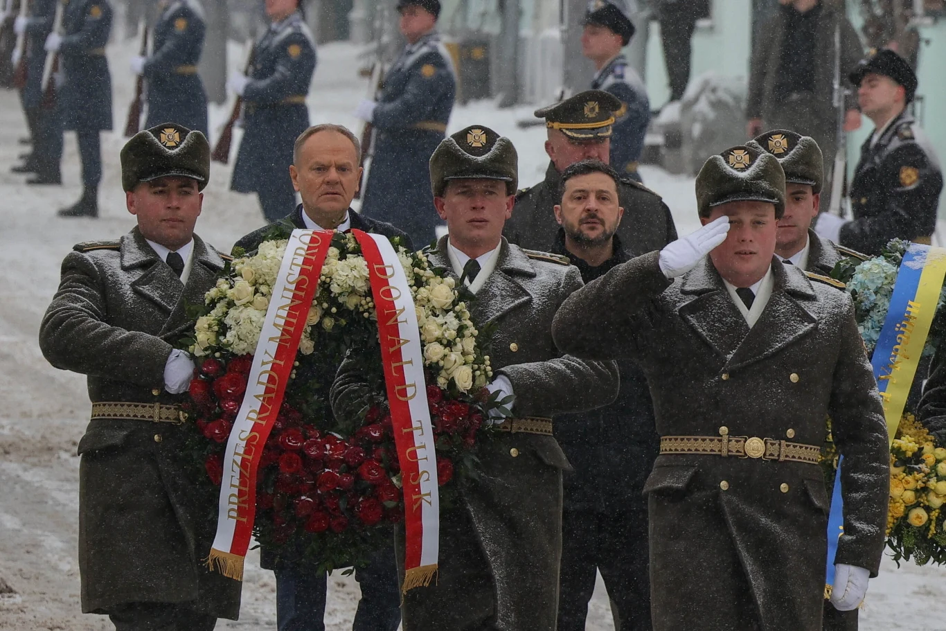 Czterech żołnierzy w szarych płaszczach niesie wiązanki kwiatów, dwaj politycy idą tuż za nimi, wokół żołnierze w niebieskich mundurach pełnią wartę podczas uroczystości na śniegu.